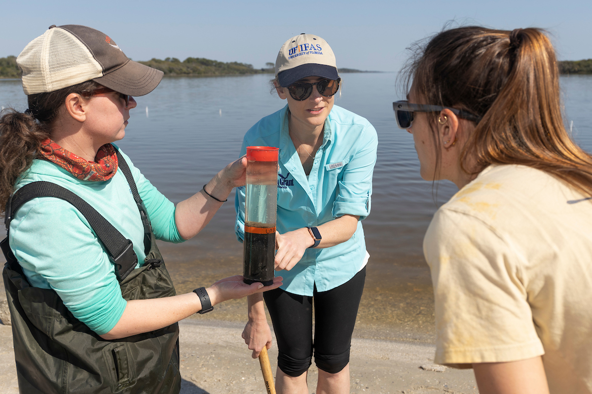 A professor stands between two graduate students on a beach, pointing to a clear cylinder containing a water and sediment core sample.