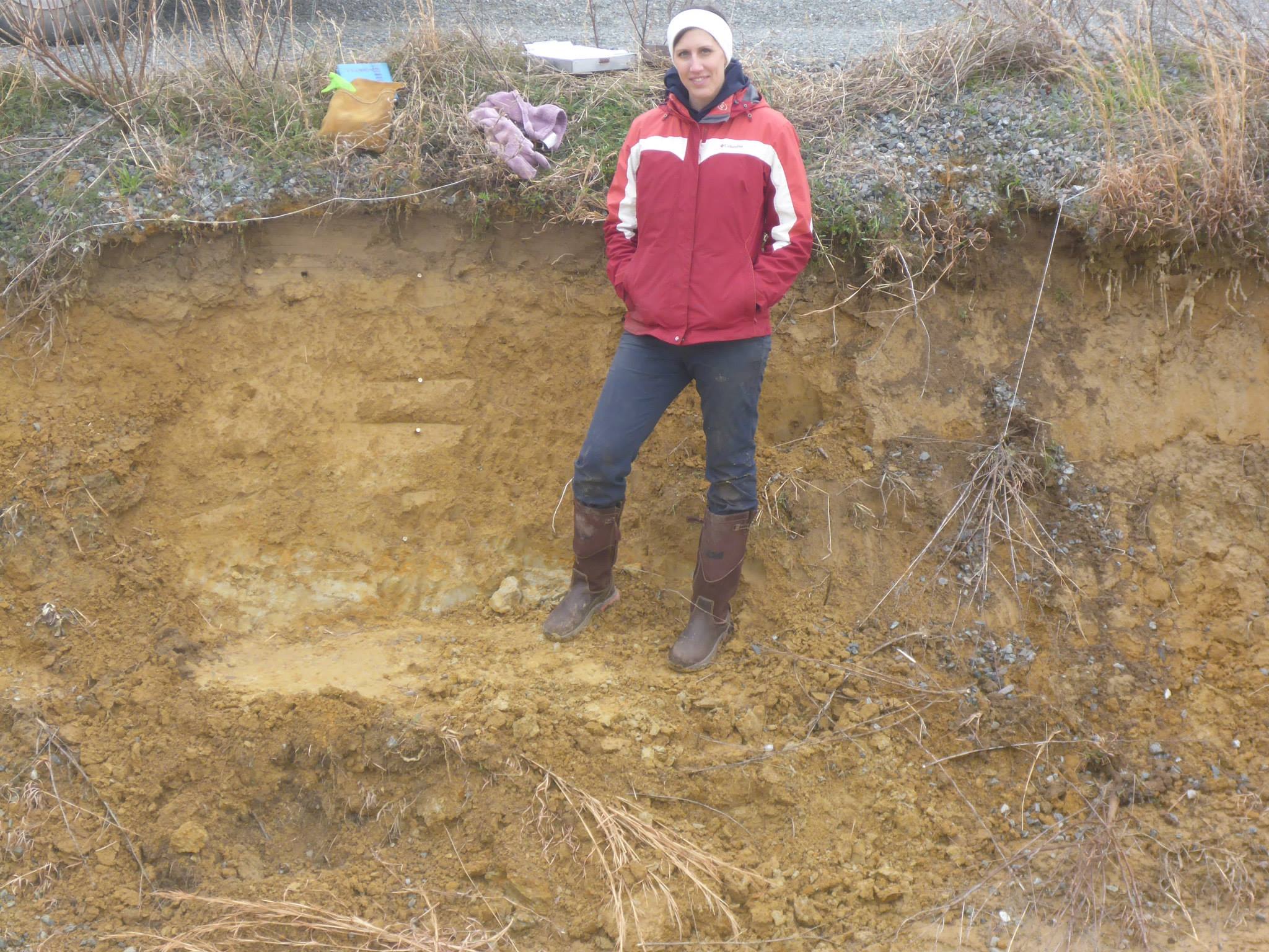 A graduate student stands in a soil pit of orange-brown soil.