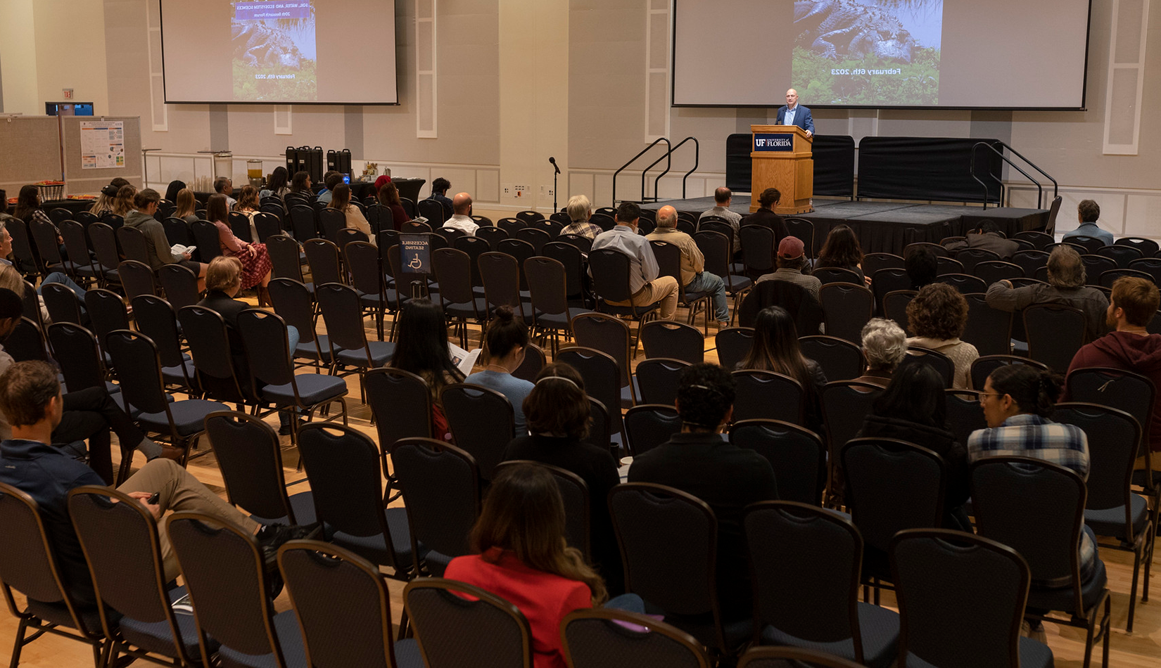 A man standing on stage, talking behind a podium to an audience at a research symposium.