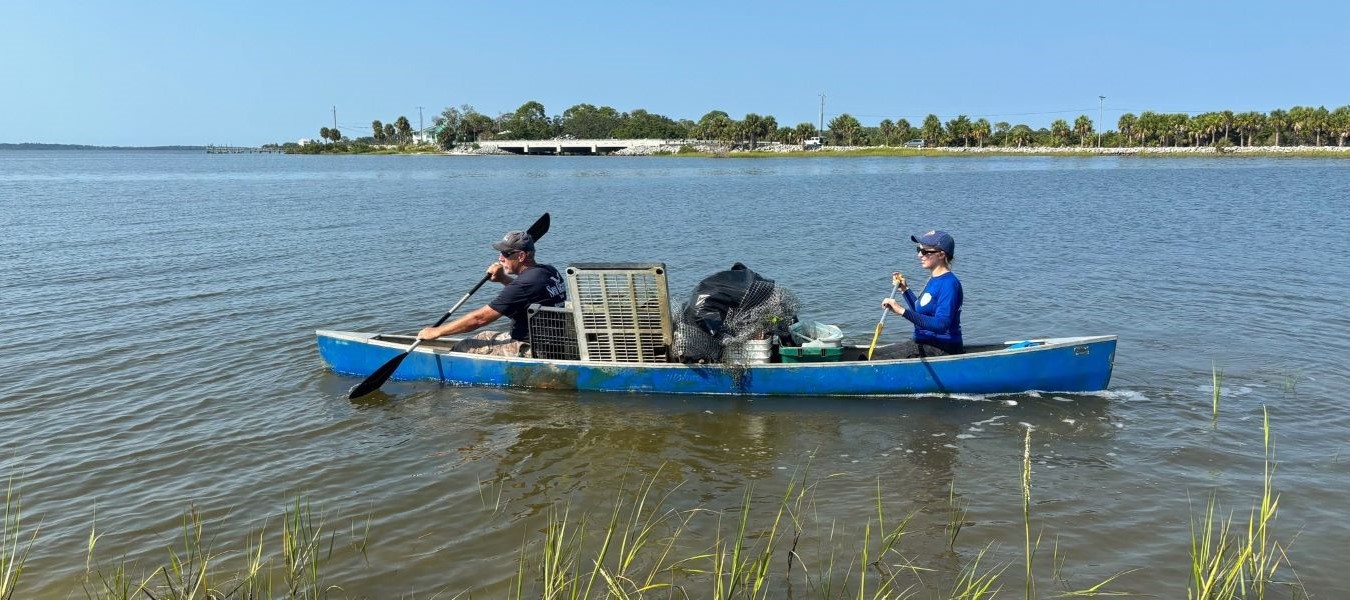 A man and woman paddle a canoe that holds beach debris in the middle.