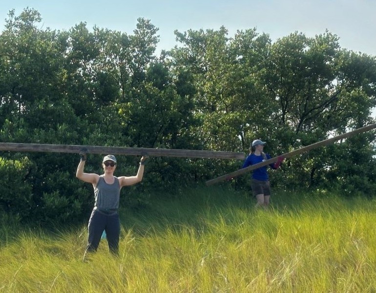 A woman stands in a clearing, holding a large wood board over her head, while another woman in the background picks up a wood board.