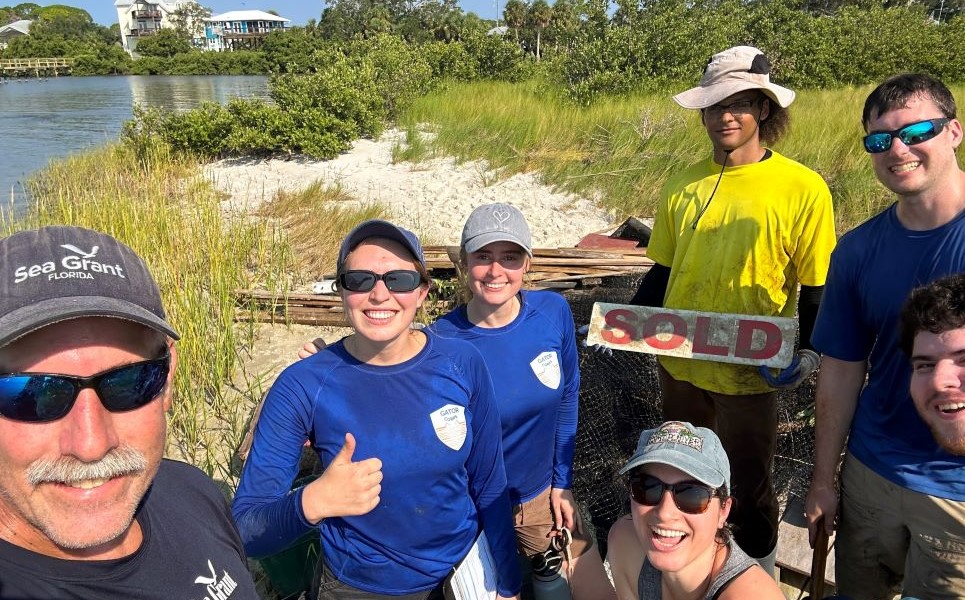 A man in sunglasses and a ballcap takes a selfie of himself and a clean up crew in front of the debris they gathered.