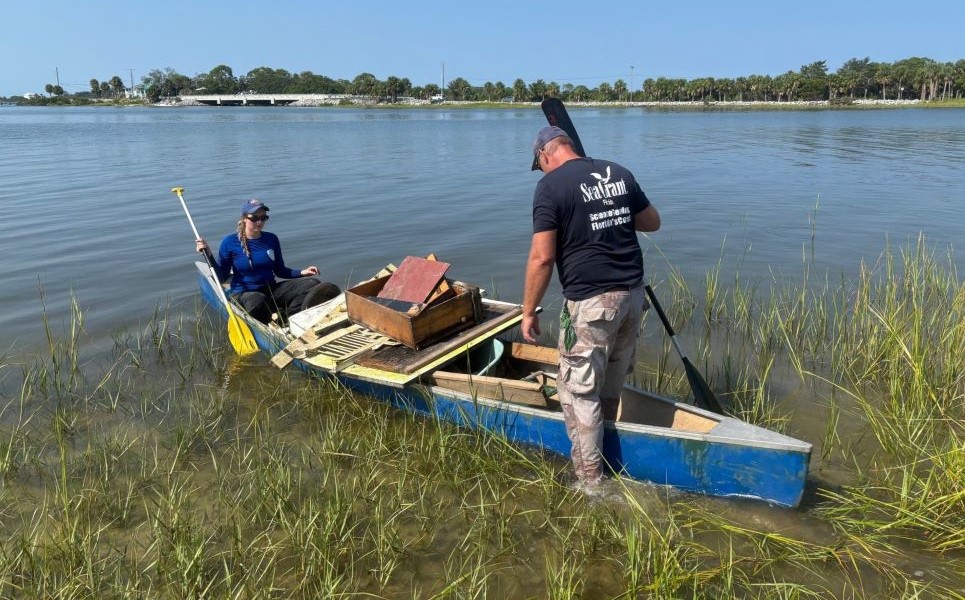 A woman sits on one side of a canoe that holds beach debris in the middle, as a man begins to sit on the other side of the canoe.