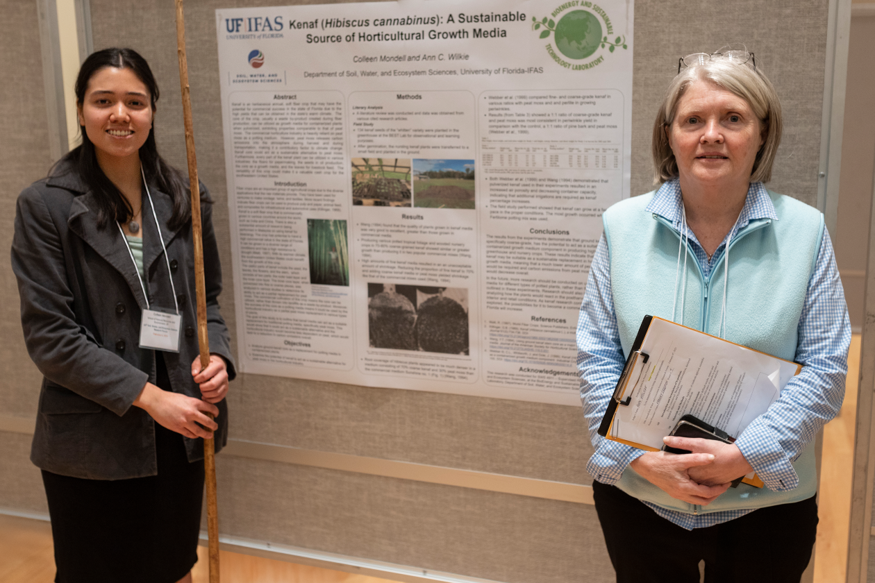 A female college student holding a long branch upright next to a research poster with her faculty advisor next to her.