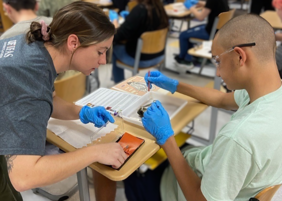 A woman stands over a student's desk, helping the student with a science experiment in the classroom.