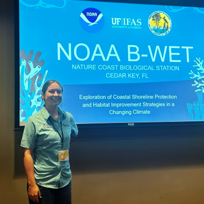 A woman stands in a darkened room in front of a large screen that has a Powerpoint presentation title slide on it.