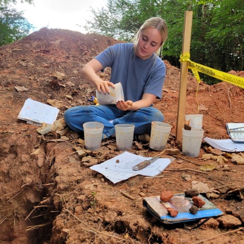 A female college student sits on red soil examining its texture as part of a soil judging competition.