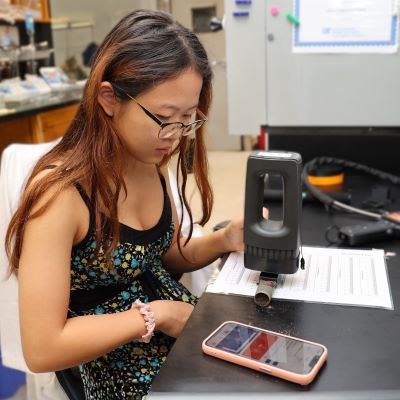 A female college student sitting at a lab bench and holding a scanning device in her left hand over a soil core sample while looking at the readings on her mobile phone app.