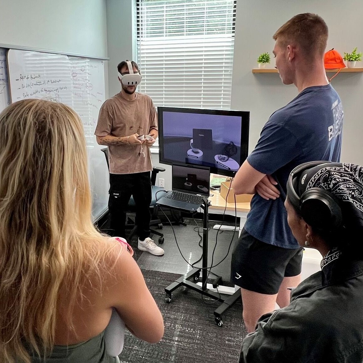 A soil science lab member demonstrates AI technology for agriculture by using a virtual reality goggle headset and hand controls as several students stand around him watching.