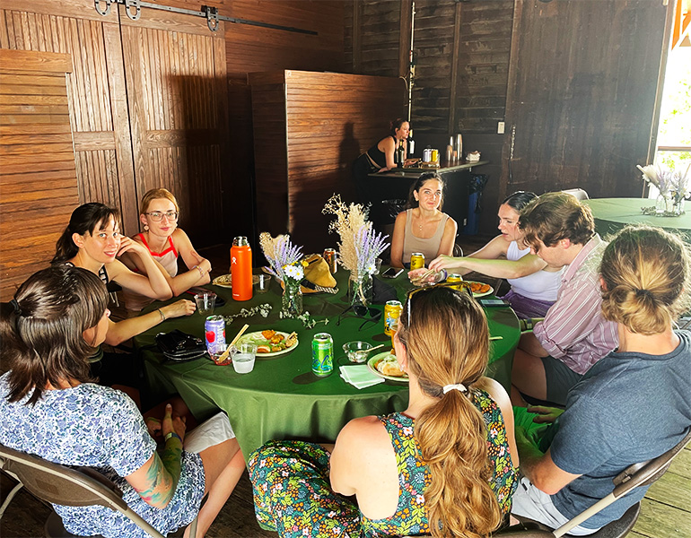 A table of 8 people sitting and eating a picnic lunch inside an old train depot.