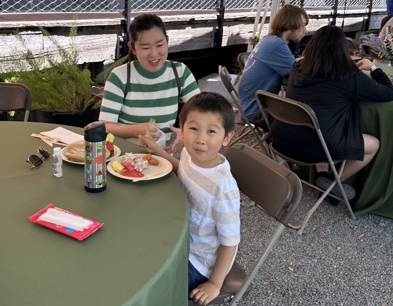 A mother and son sitting at a table under a large tent, eating a picnic lunch.