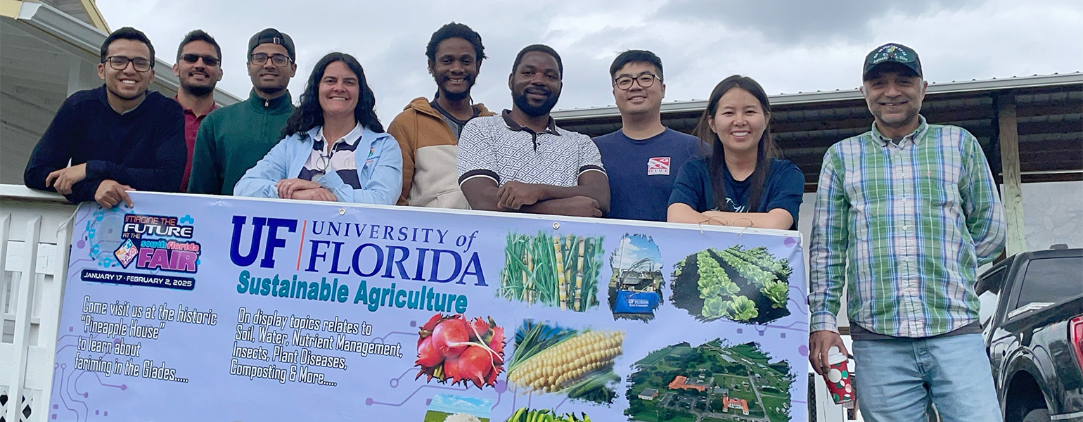 Members of a science lab team stand behind the railing of an inclined sidewalk. A banner hangs from the railing reading University of Florida with images of various vegetables.