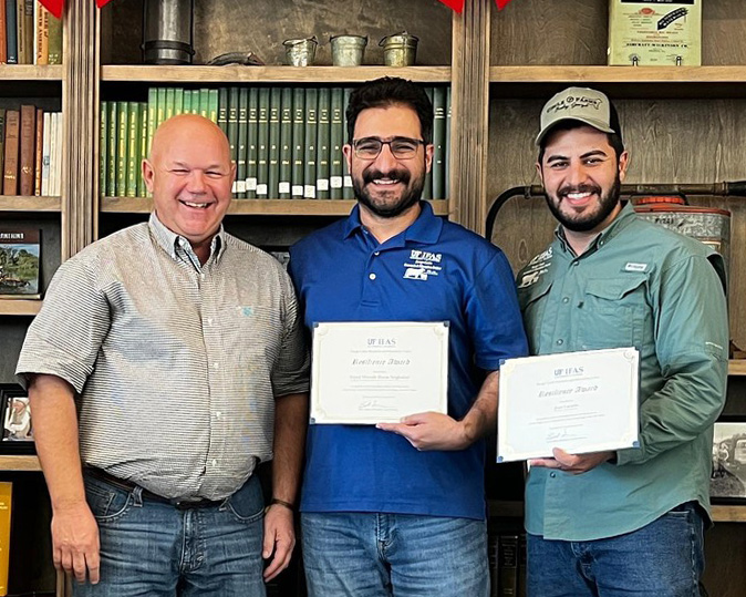 Three men stand in front of a book case with two of them holding award certificates.