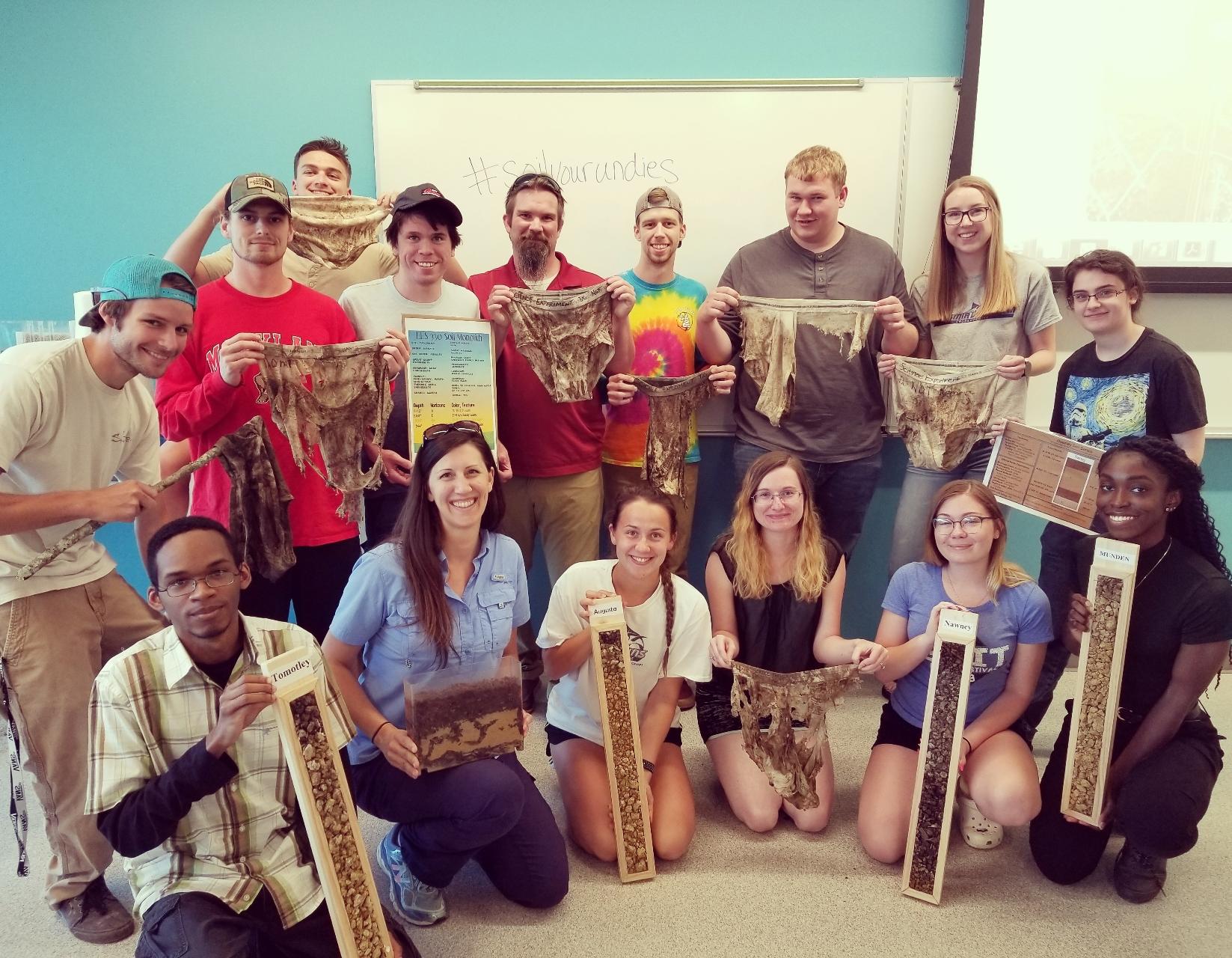 A college instructor holding a miniature soil profile in a clear case crouches in the classroom with her students for a group photo. Students in the front row hold soil profile columns. Students standing in the back row hold articles of clothing that have been degraded over time in the soil.