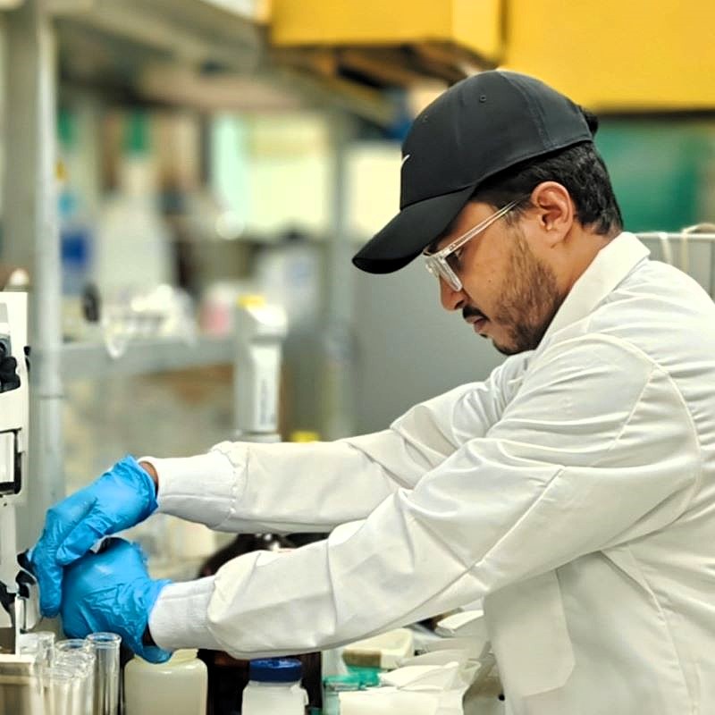 A male graduate student wearing a blue ball cap and a while lab coat with blue latex gloves works at a lab bench.