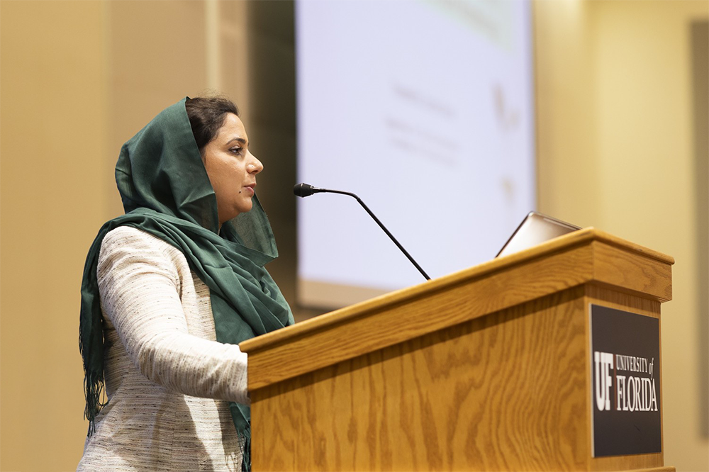 A woman wearing a headscarf stands at a podium and presents her research as a screen in the background displays her presentation.