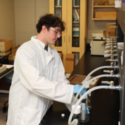 A male college student wearing a white lab coat, latex gloves and eye goggles adds a liquid to a filtered flask.