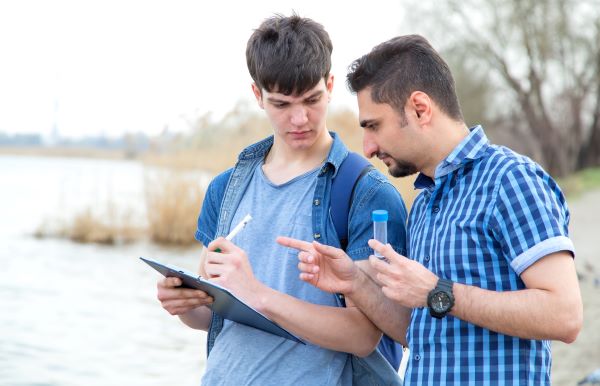 A male professor holding a vial of water talks with a college student with a clipboard about their data collection process.