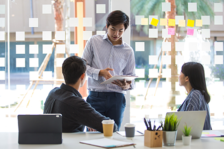 An image of a man standing in an office in front of two colleagues who are sitting. The three are discussing research collaboration.