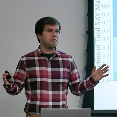 Doctoral candidate Adam Searles gives a presentation with a slide show on the screen behind him.