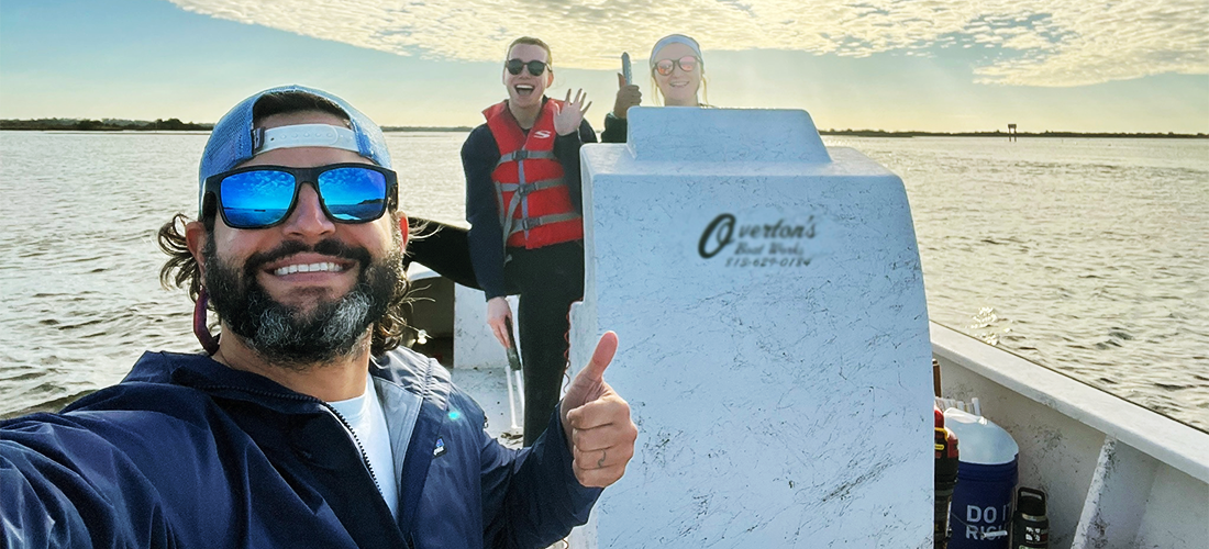 A coastal ecology professor on a small boat stands in front of two students who are in the back of the boat as all three pose for a selfie.