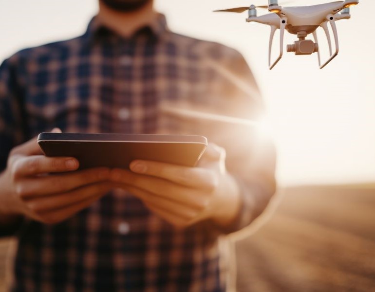 A man standing in a farm field at sunset is holding a controller to fly a drone. The drone hovers in the foreground.